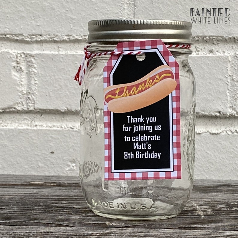 May include: A clear glass jar with a red and white checkered tag attached. The tag has a black background with the text "Thanks" and a cartoon hot dog. The text on the tag reads "Thank you for joining us to celebrate Matt's 8th Birthday".