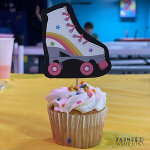 May include: A white cupcake with pink frosting and colorful sprinkles topped with a roller skate cupcake topper. The roller skate is white with a rainbow stripe and pink stars.
