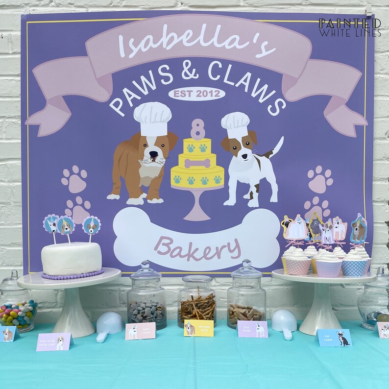 Puede incluir: Un letrero morado y rosa con el texto "Isabella's Paws & Claws Est 2012 Bakery" con un perro de dibujos animados con un gorro de cocinero y sosteniendo un hueso. Hay cupcakes con adornos con tem&aacute;tica de perros en un soporte para pasteles blanco. Hay frascos de golosinas para perros en una mesa con letreros que dicen "Dog Biscuits", "Dog Treats" y "Dog Bones".