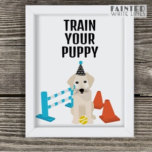May include: A white framed print with the text "TRAIN YOUR PUPPY" in black. A cartoon illustration of a yellow Labrador Retriever wearing a party hat and sitting next to a blue and white jump hurdle and orange cones. The dog is holding a yellow tennis ball.