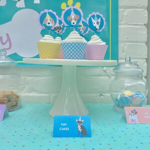 May include: A white cake stand with four cupcakes decorated with dog faces and party hats. The cupcakes are on a blue and white polka dot tablecloth. A sign reads "Pup-Cakes".