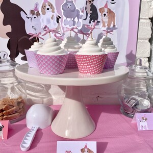 Puede incluir: Un soporte para pasteles rosa y blanco con cupcakes decorados con adornos de perros. Los cupcakes est&aacute;n etiquetados como "Pup-Cakes".