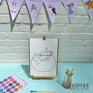 May include: A black and white line drawing of a fish wearing a party hat in a bowl. The drawing is on a white sheet of paper on an easel. There are watercolors and paintbrushes on a blue and white polka dot table cloth.