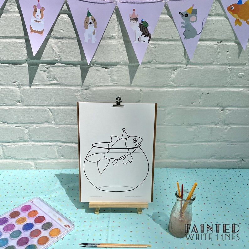 May include: A black and white line drawing of a fish wearing a party hat in a bowl. The drawing is on a white sheet of paper on an easel. There are watercolors and paintbrushes on a blue and white polka dot table cloth.