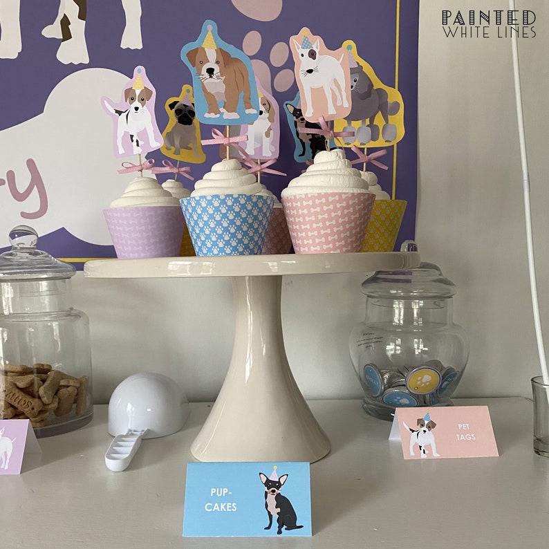 May include: A white cake stand with a variety of cupcakes decorated with dog-themed toppers. The cupcakes are on a white cake stand with a white base. The cupcakes are decorated with pink, blue, and yellow frosting. The toppers are of different breeds of dogs wearing party hats. The cupcakes are labeled "Pup Cakes" and "Pet Tags".