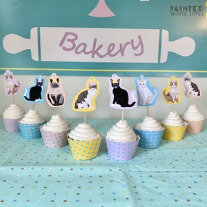 Puede incluir: Una mesa con un mantel azul claro con lunares blancos. Hay 8 cupcakes con glaseado blanco y forros de cupcakes con lunares de diferentes colores. Cada cupcake tiene un adorno de gato con un gorro de fiesta. El letrero sobre los cupcakes dice "Bakery" con un rodillo.