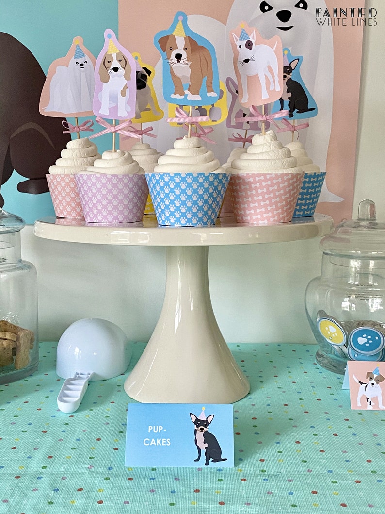 May include: A display of cupcakes with dog-themed toppers, presented on a white cake stand. The cupcakes have paw print-patterned liners in pink, blue, and purple. A sign reads "PUP-CAKES" with a dog illustration.