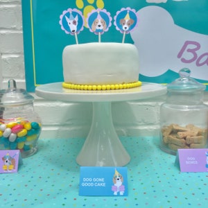 Pode incluir: Um bolo branco com cobertura amarela e decora&ccedil;&otilde;es com tema de c&atilde;es em um suporte de bolo branco. O bolo est&aacute; em uma mesa com uma toalha de mesa azul e branca de bolinhas. H&aacute; dois potes com guloseimas na mesa, um com doces coloridos e o outro com biscoitos para c&atilde;es. H&aacute; tamb&eacute;m duas placas na mesa, uma que diz "Dog Gone Good Cake" e a outra que diz "Dog Bones".
