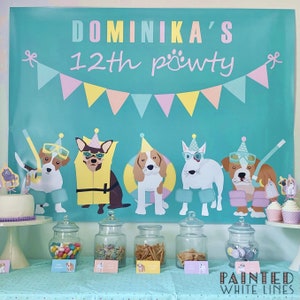 May include: A turquoise blue party backdrop with the text "Dominika's 12th pawty" and a string of pastel pink, yellow, and blue flags. Five cartoon dogs wearing party hats and goggles are on the backdrop. A table with a white cake, cupcakes, and glass jars filled with candy and snacks is in front of the backdrop.