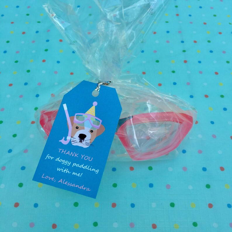 May include: A blue and white thank you tag with a cartoon dog wearing a party hat and goggles. The tag says "Thank you for doggy paddling with me! Love, Alessandra". The tag is attached to a clear plastic bag with pink swimming goggles inside.