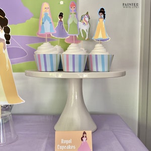 May include: Three cupcakes with white frosting and colorful striped cupcake liners on a white cake stand. The cupcakes are topped with princess character toppers. A sign in front of the stand reads "Royal Cupcakes".