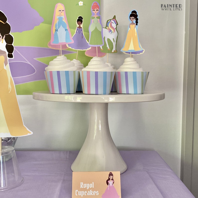 May include: Three cupcakes with white frosting and colorful striped cupcake liners on a white cake stand. The cupcakes are topped with princess character toppers. A sign in front of the stand reads "Royal Cupcakes".
