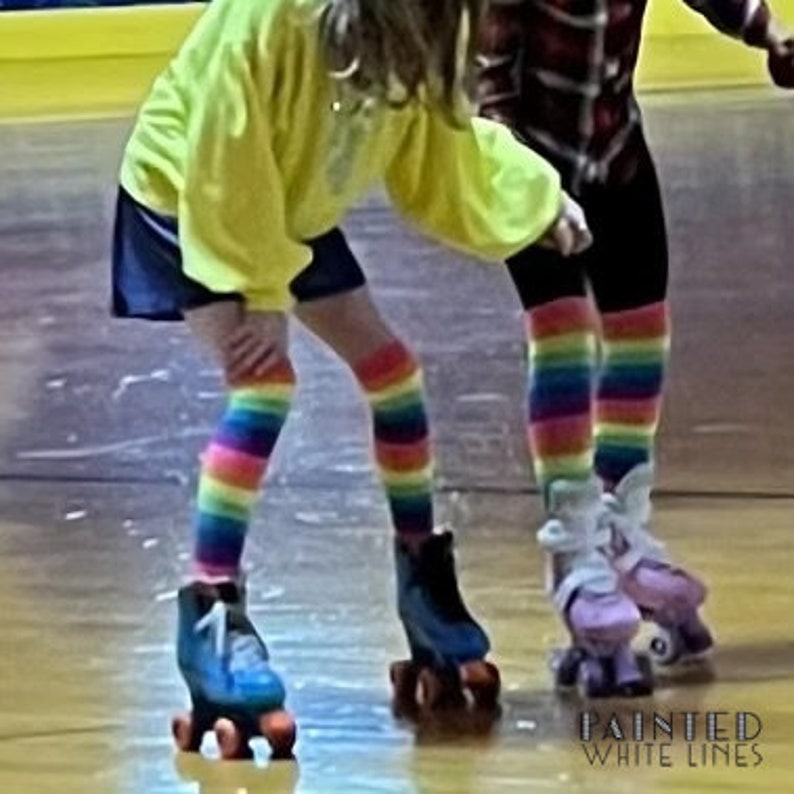 May include: Two people wearing rainbow striped socks and roller skates on a wooden floor. The person in the foreground is wearing a yellow shirt and a blue skirt. The person in the background is wearing a red and black plaid shirt.