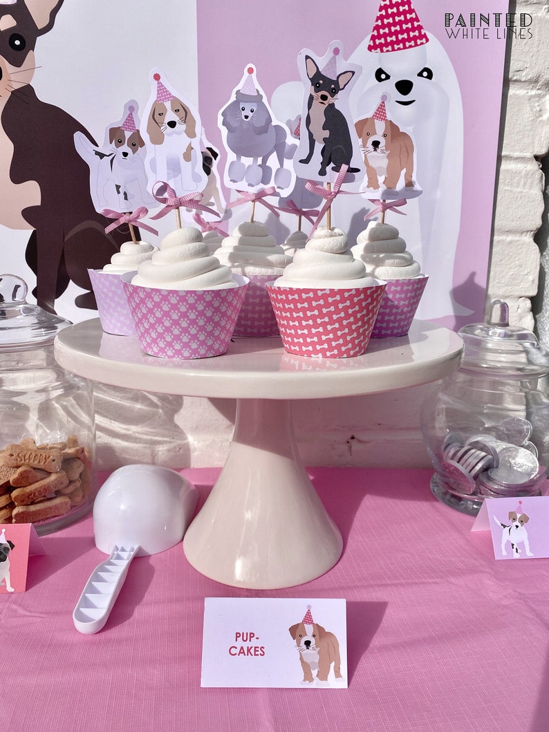 May include: A pink and white cake stand with cupcakes decorated with dog toppers. The cupcakes are labeled "Pup-Cakes".