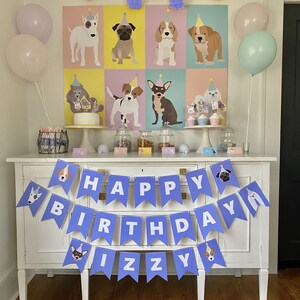 May include: A purple and white 'Happy Birthday Izzy' banner with dog illustrations hangs above a white buffet table decorated with a dog-themed party. The table is set with a cake, cupcakes, and other treats. There are balloons and a poster with dog illustrations in the background.