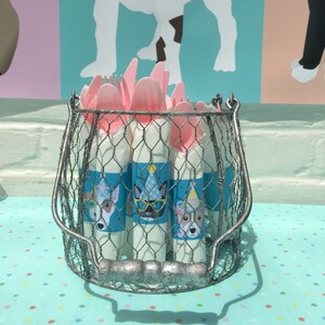 May include: A wire basket filled with pink plastic forks and rolled napkins. The napkins are decorated with illustrations of dogs wearing party hats and glasses. The basket sits on a light blue surface with colorful polka dots.