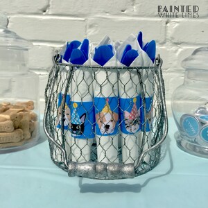 May include: A silver wire basket filled with white paper cones decorated with blue and white dog faces. The cones are topped with blue plastic forks. The basket is sitting on a light blue table.