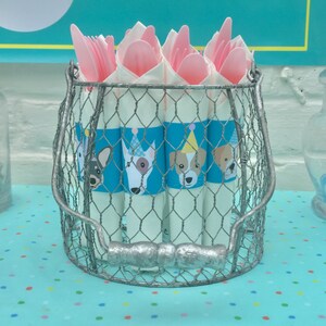 May include: A silver wire basket filled with white paper cones decorated with blue and white dog illustrations. The cones are topped with pink plastic forks and spoons.
