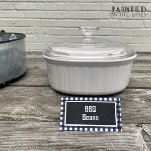 May include: White ceramic casserole dish with a glass lid, filled with BBQ beans. The dish is sitting on a wooden table with a black and white checkered sign that reads "BBQ Beans".