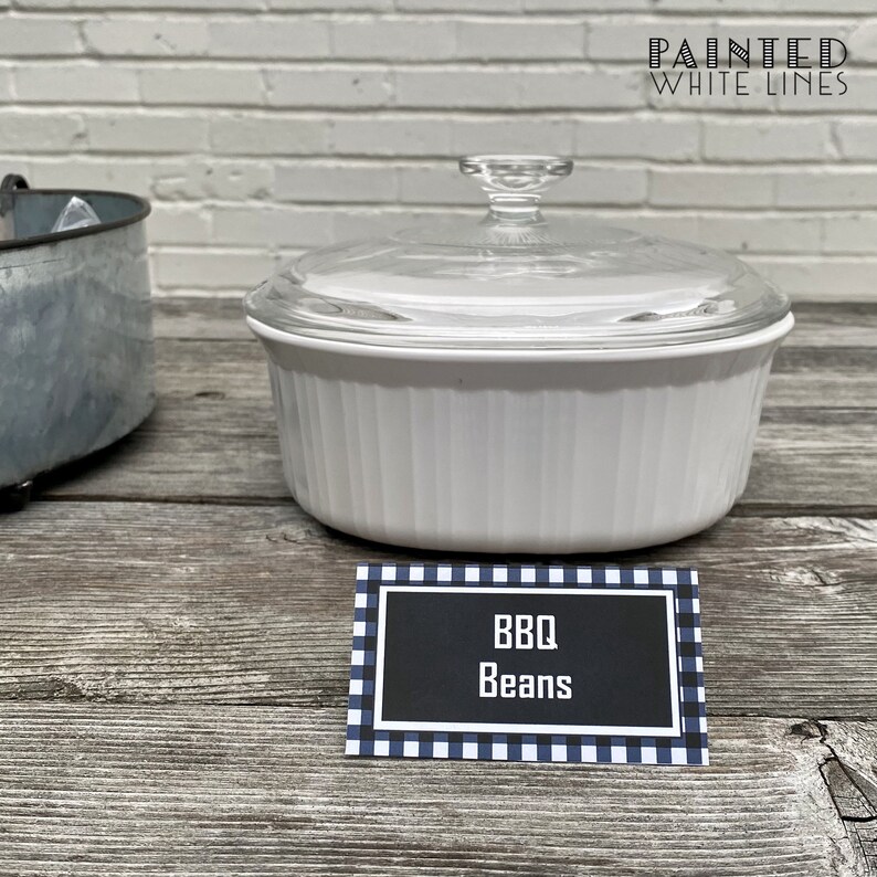 May include: White ceramic casserole dish with a glass lid, filled with BBQ beans. The dish is sitting on a wooden table with a black and white checkered sign that reads "BBQ Beans".