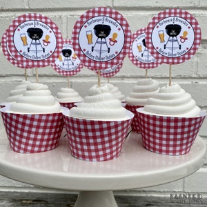 Pode incluir: Forminhas de cupcake em gingham vermelho e branco com cobertura branca e um topo de cupcake redondo de papel branco com uma ilustra&ccedil;&atilde;o em preto e branco de uma grelha, cerveja e uma chupeta. O texto no topo diz "Barbeque & Brewskis The Smiths * Before Diaper Duties *"