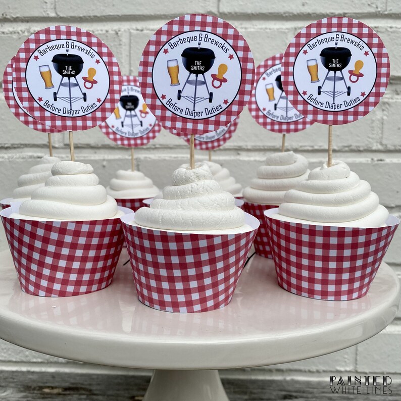 Pode incluir: Forminhas de cupcake em gingham vermelho e branco com cobertura branca e um topo de cupcake redondo de papel branco com uma ilustra&ccedil;&atilde;o em preto e branco de uma grelha, cerveja e uma chupeta. O texto no topo diz "Barbeque & Brewskis The Smiths * Before Diaper Duties *"