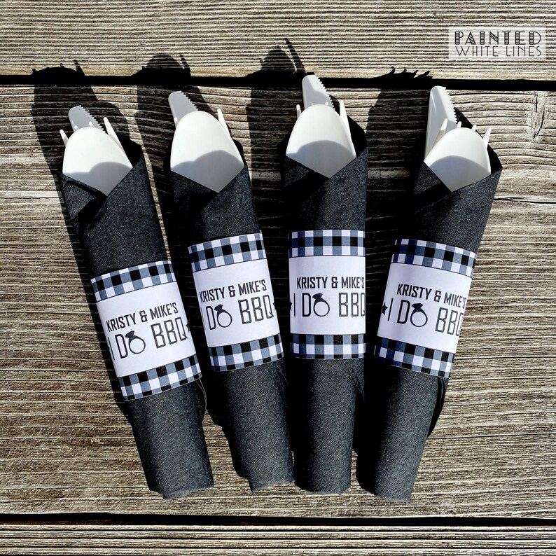 May include: Four black napkins with white and black checkered paper rings that say "Kristy & Mike's I Do BBQ" with a ring icon. The napkins are wrapped around white plastic utensils.