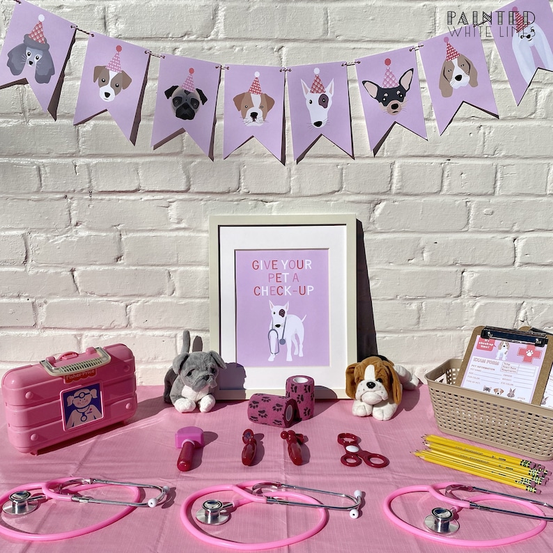 May include: A pink table set up for a pretend pet doctor's office. There are two pink toy stethoscopes, a toy medical kit, a toy hammer, bandages, and two stuffed dogs. A framed sign reads "Give your pet a check-up".