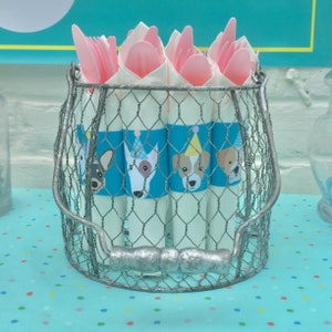 Op de afbeelding: Een zilveren draadmand gevuld met witte papieren kegels versierd met blauwe en witte hondenillustraties. De kegels zijn bedekt met roze plastic vorken en lepels.