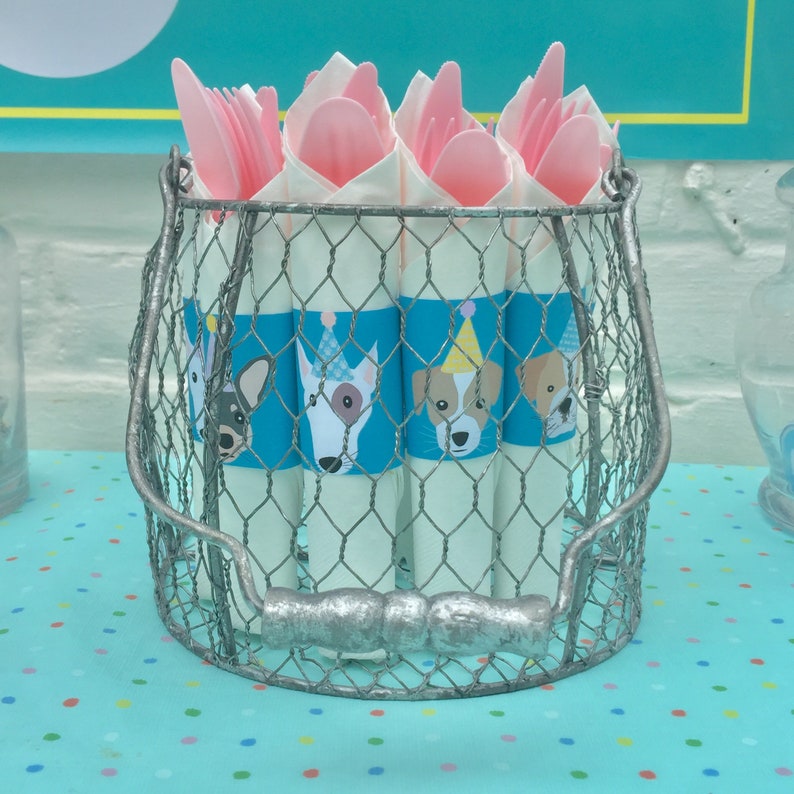 Op de afbeelding: Een zilveren draadmand gevuld met witte papieren kegels versierd met blauwe en witte hondenillustraties. De kegels zijn bedekt met roze plastic vorken en lepels.