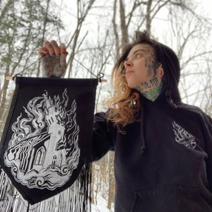 Könnte beinhalten: Eine schwarze Wandbehang aus Stoff mit einer weißen Illustration einer Kirche, die in Flammen steht. Der Wandbehang hat einen Fransenrand und hängt an einer Holzleiste.