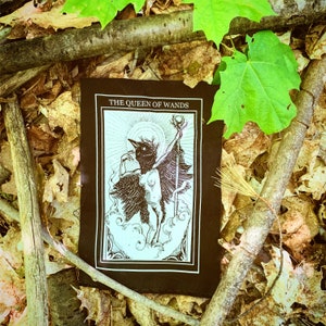 May include: A black tarot card with the words "The Queen of Wands" printed on it. The card features a black and white illustration of a bird-like creature with wings outstretched, holding a staff with a star at the top.