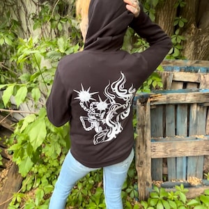 May include: Black hooded sweatshirt with a white graphic design on the back. The design features a stylized illustration with chain and flame elements. The person is wearing light blue jeans and white socks.