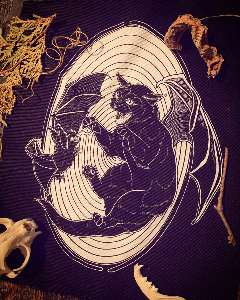 May include: Black and white illustration of a cat with bat wings, fighting a bat. The image is on a dark fabric background.