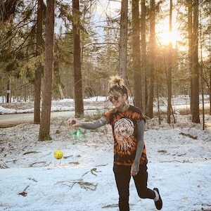 May include: A person wearing a tie-dye shirt with a skull and floral design print, standing in a snowy woodland. They are wearing sunglasses and a nose ring, and holding a yellow ball.