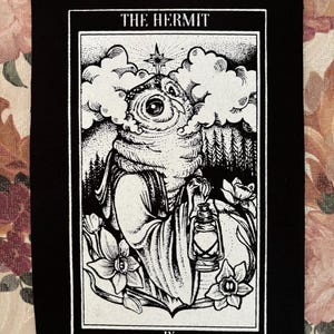 K&ouml;nnte beinhalten: Schwarz-wei&szlig; gestickter Patch mit der Tarotkarte Der Einsiedler. Die Karte zeigt eine Person mit Kapuze, die eine Laterne h&auml;lt und in einem Wald steht. Die Kartennummer IX befindet sich am unteren Rand der Karte.