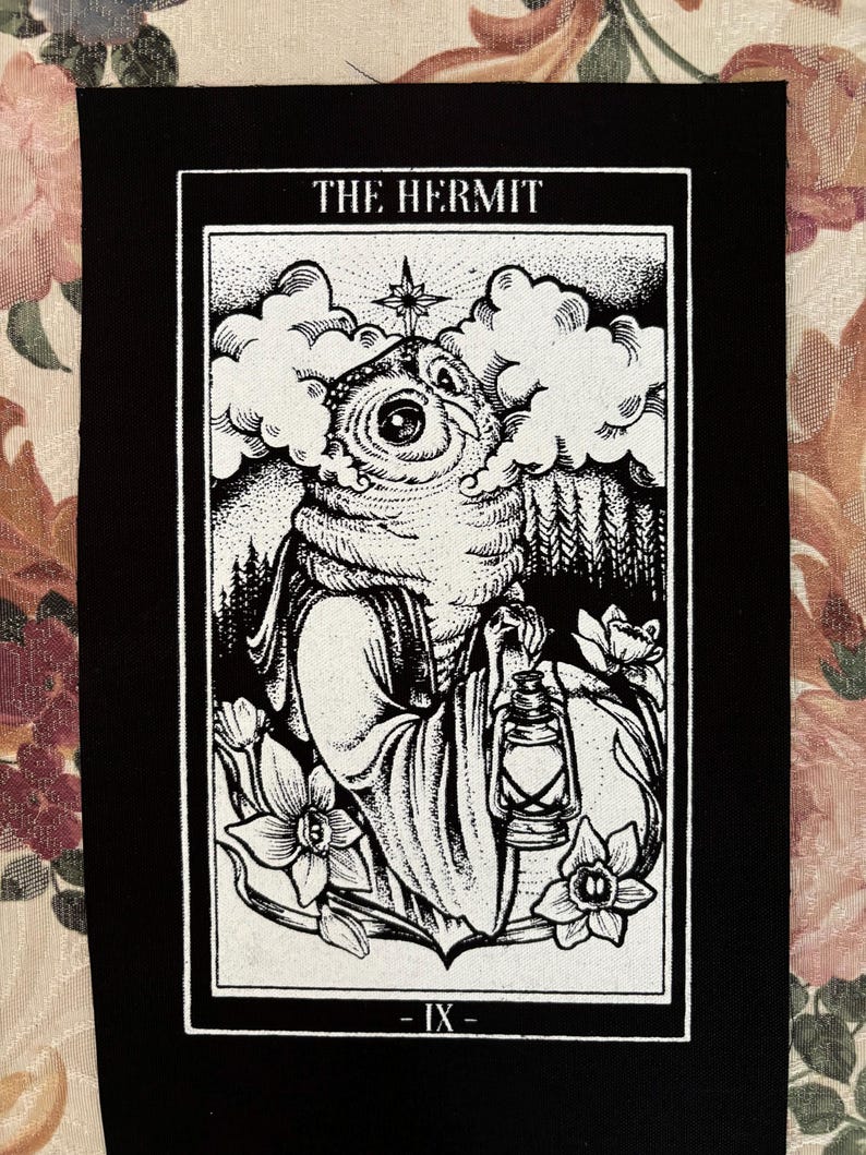 K&ouml;nnte beinhalten: Schwarz-wei&szlig; gestickter Patch mit der Tarotkarte Der Einsiedler. Die Karte zeigt eine Person mit Kapuze, die eine Laterne h&auml;lt und in einem Wald steht. Die Kartennummer IX befindet sich am unteren Rand der Karte.
