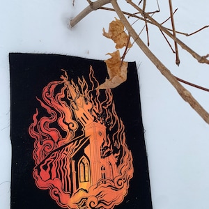 Pode incluir: Um remendo preto com uma igreja em chamas. As chamas são laranja e amarelas, e a igreja é delineada em preto. O remendo está sobre um fundo branco.