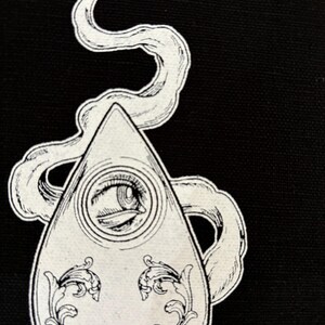 Puede incluir: Una ilustraci&oacute;n en blanco y negro de una tabla Ouija con un solo ojo en el centro y detalles ornamentados alrededor de los bordes. La tabla est&aacute; rodeada por un dise&ntilde;o de humo en espiral.