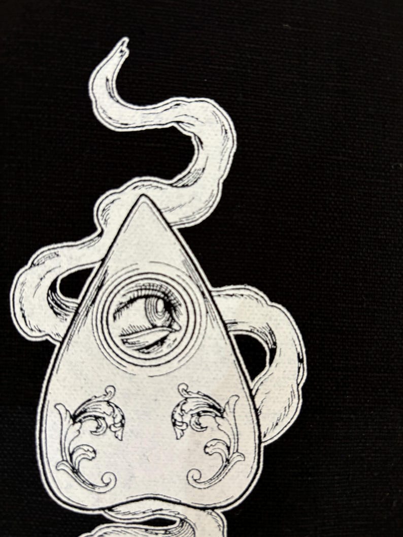 Puede incluir: Una ilustraci&oacute;n en blanco y negro de una tabla Ouija con un solo ojo en el centro y detalles ornamentados alrededor de los bordes. La tabla est&aacute; rodeada por un dise&ntilde;o de humo en espiral.
