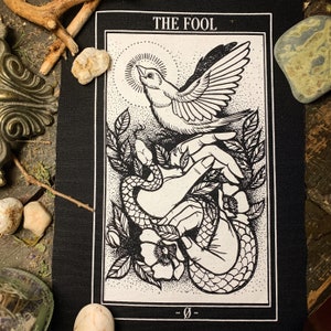 May include: Black and white tarot card patch with the text "THE FOOL" at the top. The card depicts a hand holding a bird with a snake wrapped around it. The hand is surrounded by flowers and leaves.