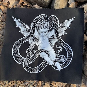 May include: A black and white embroidered patch featuring a cherub with wings, wrapped in a snake. The cherub has a devilish expression and is surrounded by a snake with a detailed pattern.