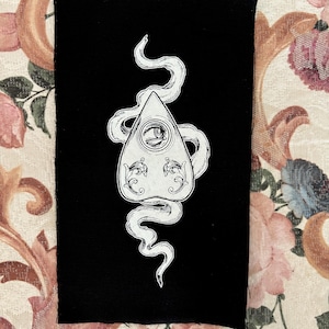 Puede incluir: Un parche de tela negro con un contorno blanco de una tabla Ouija con una serpiente envuelta alrededor.