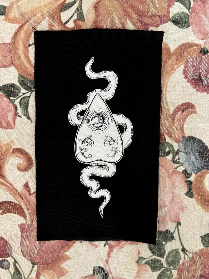 Puede incluir: Un parche de tela negro con un contorno blanco de una tabla Ouija con una serpiente envuelta alrededor.