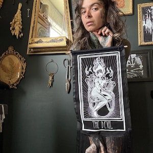 May include: A black tapestry featuring a white illustration of the Devil tarot card. The card depicts a nude figure with horns and wings, chained to a pedestal. The text "THE DEVIL" is printed beneath the image.