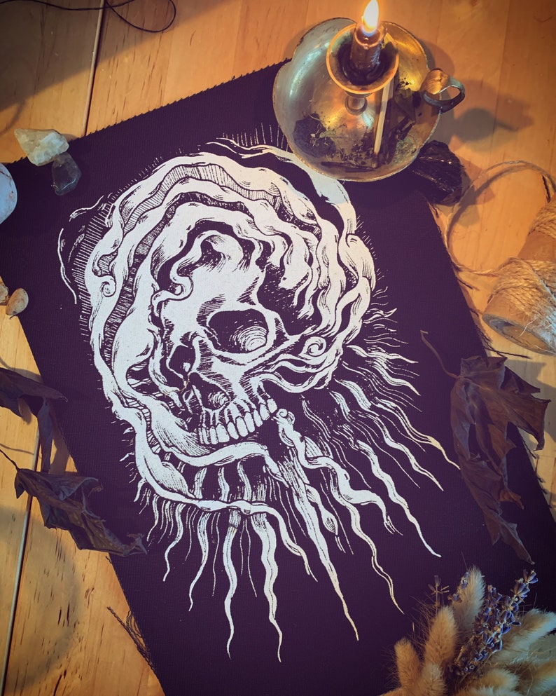 Op de afbeelding: Een zwarte stoffen patch met een wit schedelontwerp. De schedel is omgeven door draaiende lijnen en heeft &eacute;&eacute;n oog. De patch is perfect om een ​​gotische touch aan elk project toe te voegen.