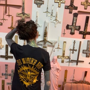 May include: A person wearing a black t-shirt with a gold graphic of a butterfly and the text "The Witch's Nest". The person is standing in front of a wall decorated with numerous inverted crosses.