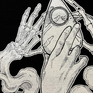 May include: A black and white illustration of a skeletal hand holding a white ouija board with a ghostly hand reaching out from the smoke below.