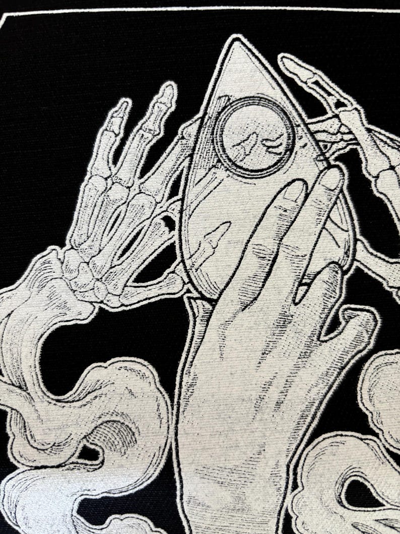 May include: A black and white illustration of a skeletal hand holding a white ouija board with a ghostly hand reaching out from the smoke below.