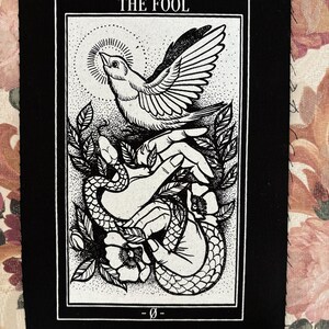 Op de afbeelding: Zwart-witte tarotkaart patch met de tekst "THE FOOL" bovenaan. De kaart toont een hand die een vogel en een slang vasthoudt met een bloem op de achtergrond.
